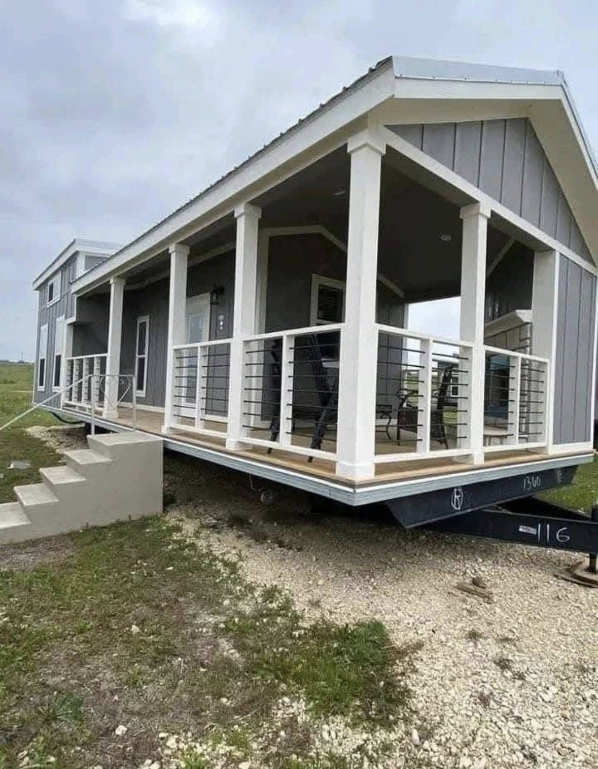 Modern tiny house on wheels with a porch and railing sits on a grassy landscape. The neutral tones and overcast sky create a serene, minimalist vibe.