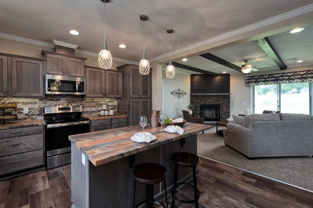 Modern kitchen and living area featuring dark wood cabinets, stainless steel appliances, a rustic island with bar stools, elegant pendant lights, and a cozy gray living room set with a fireplace.