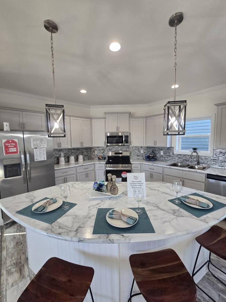Modern kitchen featuring a marble island with teal placemats and wooden stools, pendant lights, stainless steel appliances, and gray cabinetry.