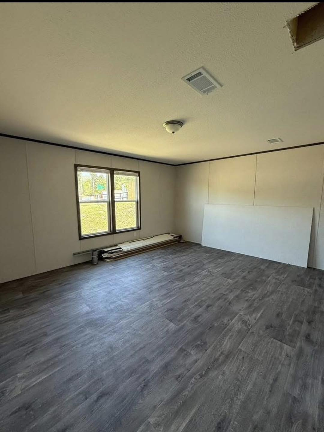 An empty room with light beige walls and dark wood laminate flooring. Sunlight streams through two windows. A board and rolls of paper lie by the wall.