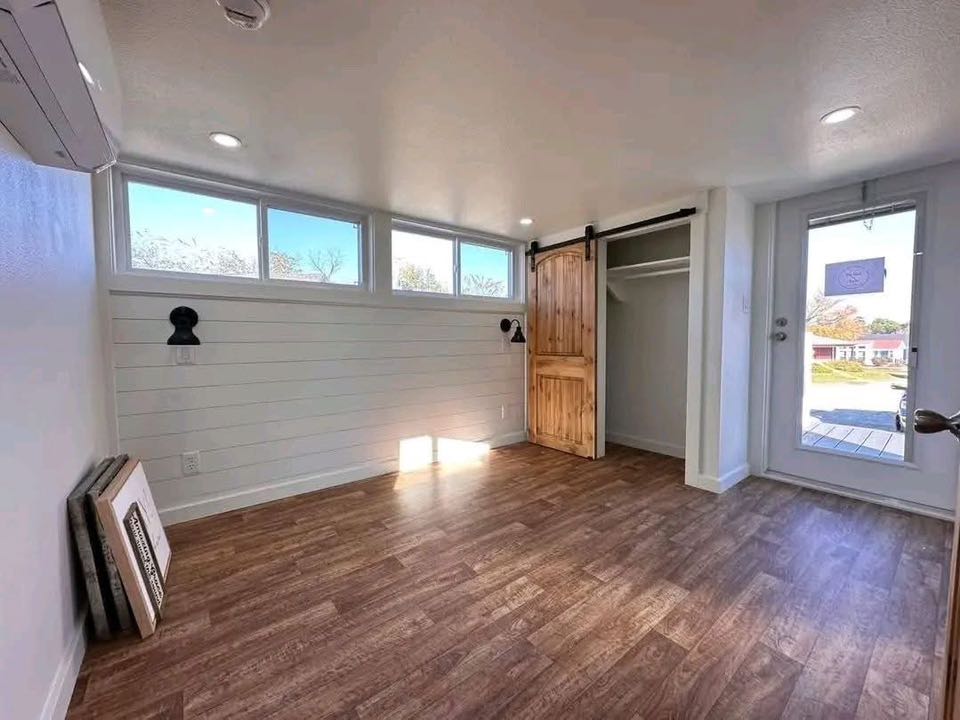 Bright, empty room with wood floor, white shiplap walls, and four small windows. Features a wooden sliding barn door and a glass door leading outside.