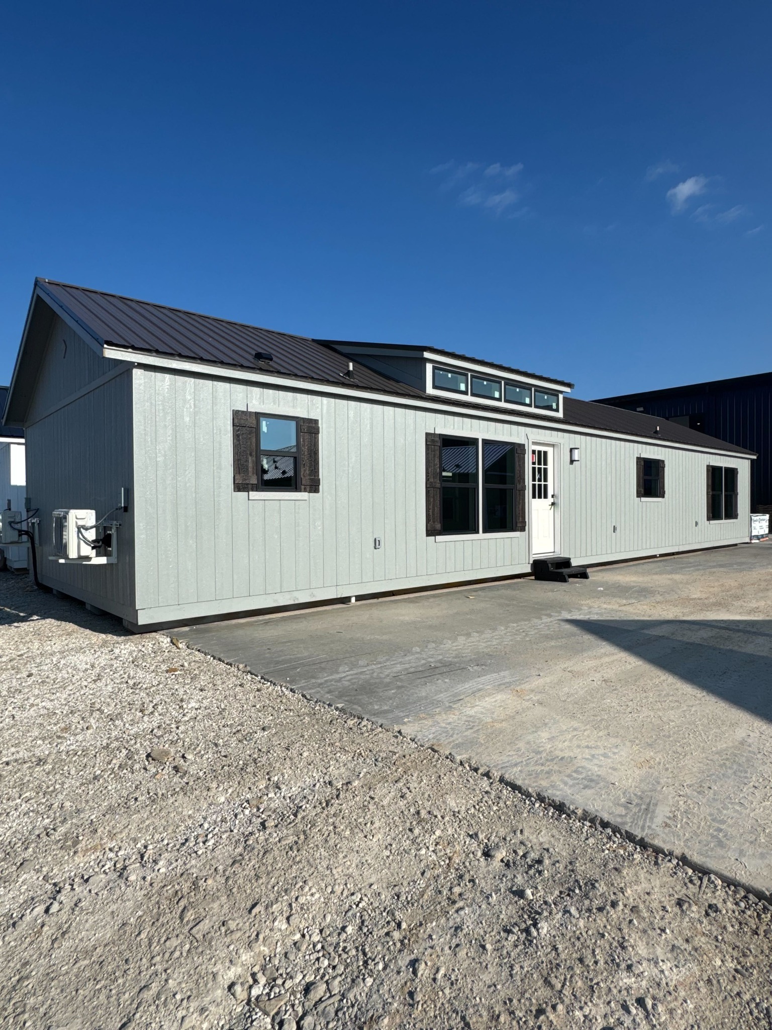 Modern tiny house with a light gray exterior and black roof, featuring large windows and a white door, stands on a concrete pad under a clear blue sky.
