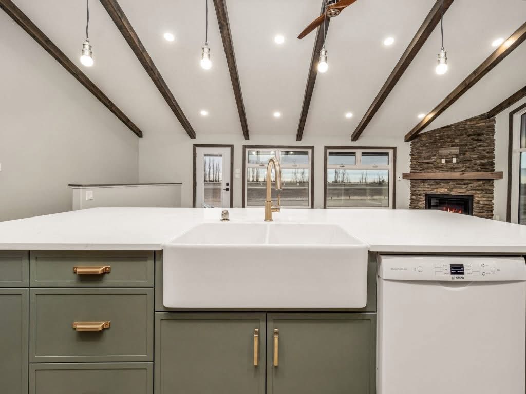 Modern kitchen with green cabinets, a large white sink, and gold faucet. Exposed beams and hanging lights on the ceiling, stone fireplace in the background. Cozy and inviting.