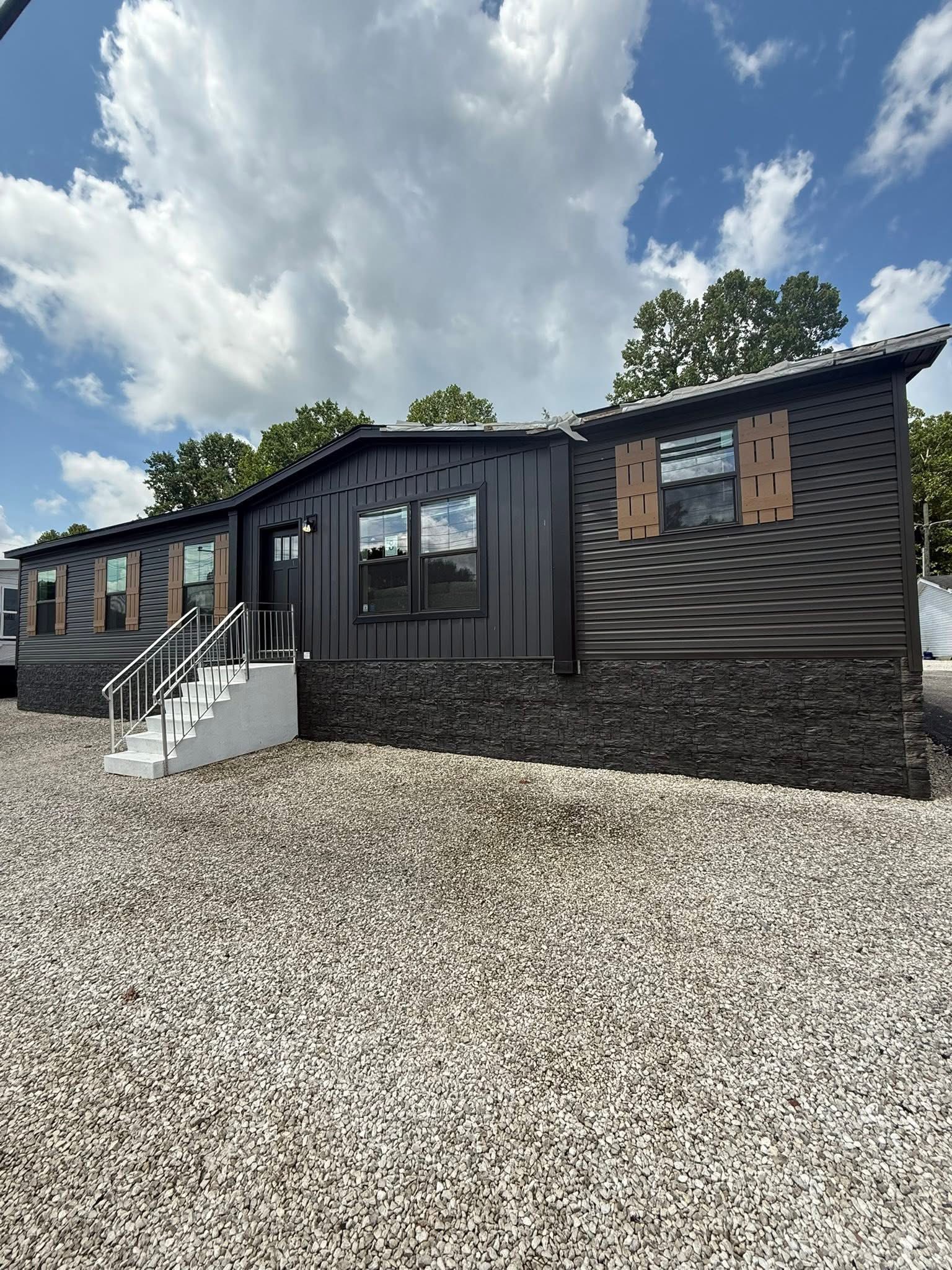 Modern black mobile home with white steps and railing, set on gravel. The sky is partly cloudy, and trees are visible in the background.