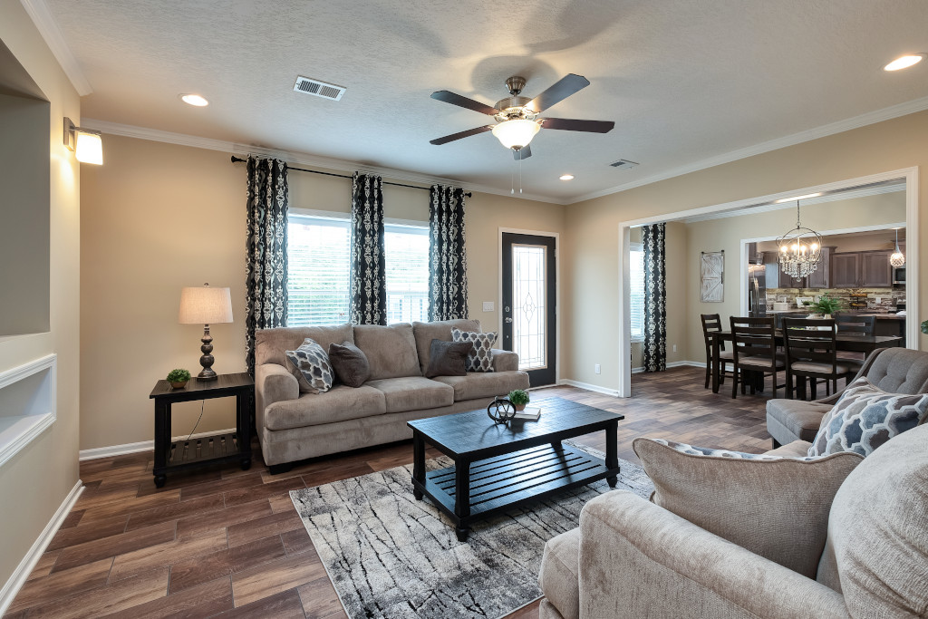 A cozy living room with beige sofas and patterned cushions, a black coffee table, and a rug. Warm lighting and dark curtains create an inviting ambiance. Dining area visible.