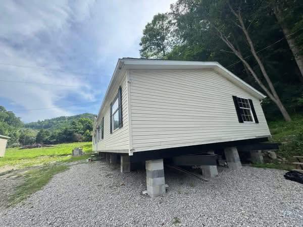 A beige mobile home is elevated on concrete blocks, surrounded by gravel, with trees and a grassy landscape in the background under a partly cloudy sky.