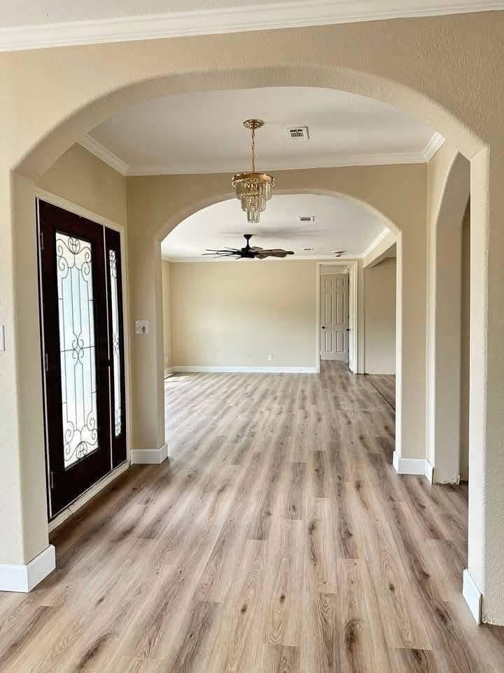 Spacious hallway with light wood flooring, arched doorways, a chandelier, and a ceiling fan. Natural light from a decorative glass door creates a welcoming ambiance.