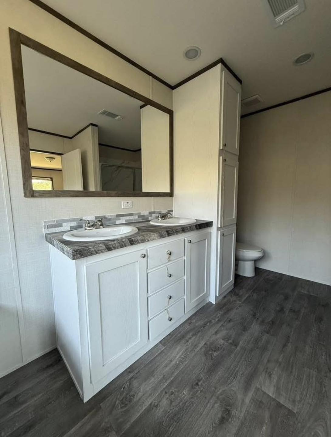 Spacious bathroom with dual sinks, marble countertop, and a large rectangular mirror. Dark wood flooring complements white cabinetry, creating a modern feel.