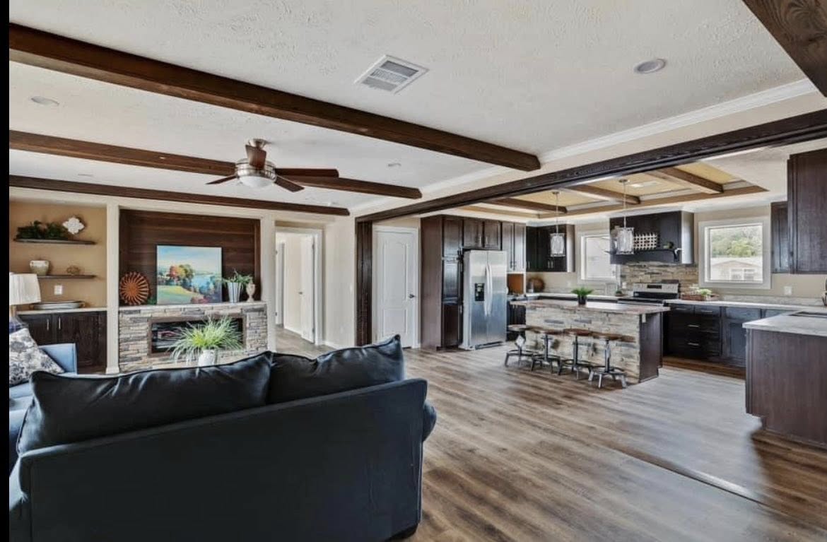 Stylish open-concept living area with wooden beams, modern kitchen, cozy fireplace, and dark wood furniture. Warm and inviting atmosphere.