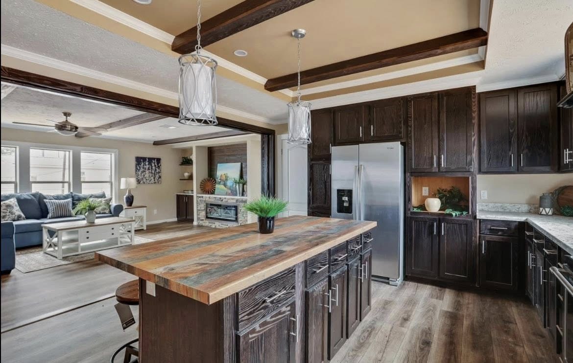 Spacious kitchen with wooden beams, dark cabinets, and a large island with a striped wood countertop. It opens to a cozy living room with a blue sofa.