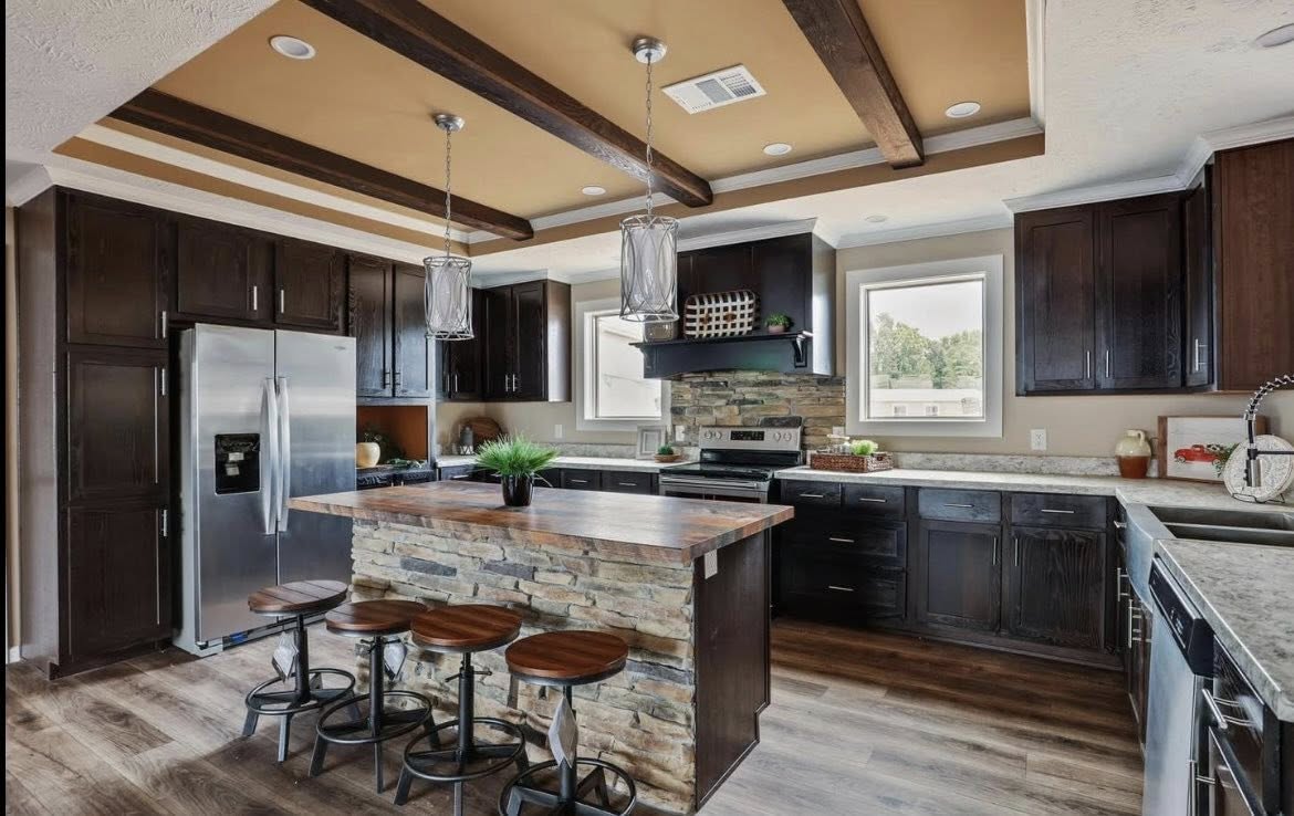 Modern kitchen with dark wooden cabinets, a stone island, and barstools. Stainless steel appliances and exposed ceiling beams add a sleek touch. Natural light through windows enhances the warm, inviting atmosphere.