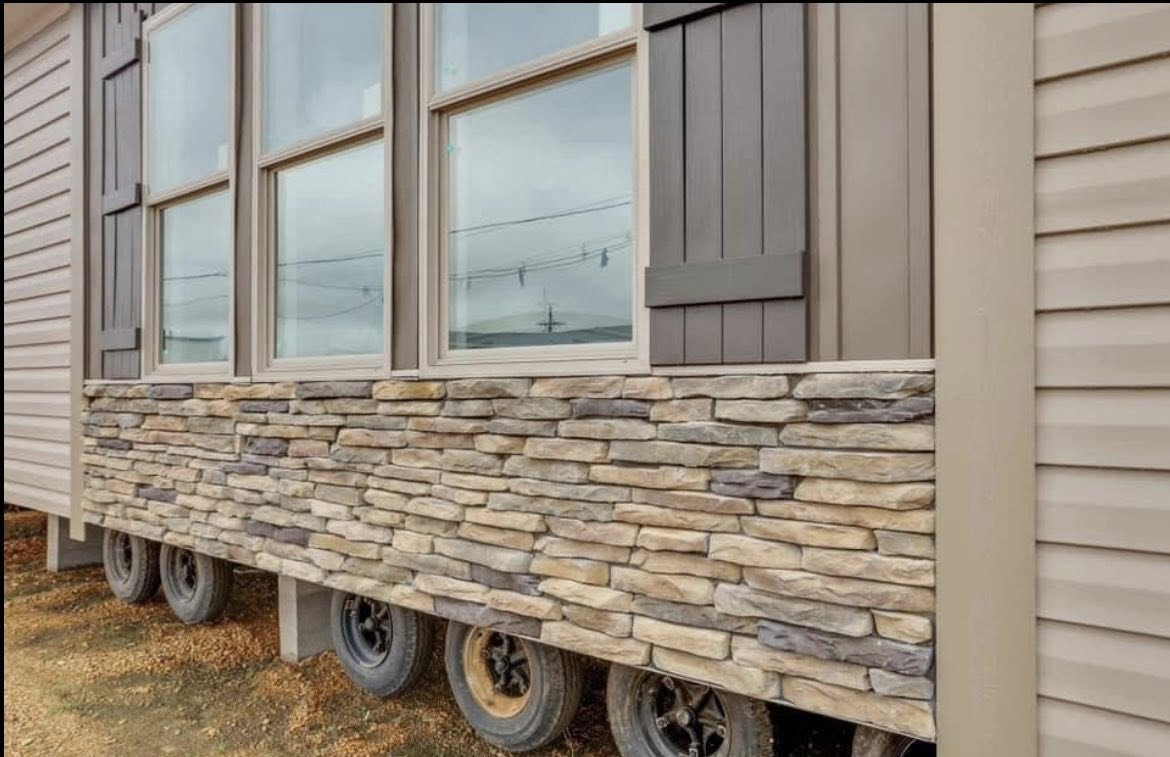 Close-up of a mobile home exterior with stone veneer at the bottom and beige siding. Multiple wheels visible underneath, suggesting mobility.