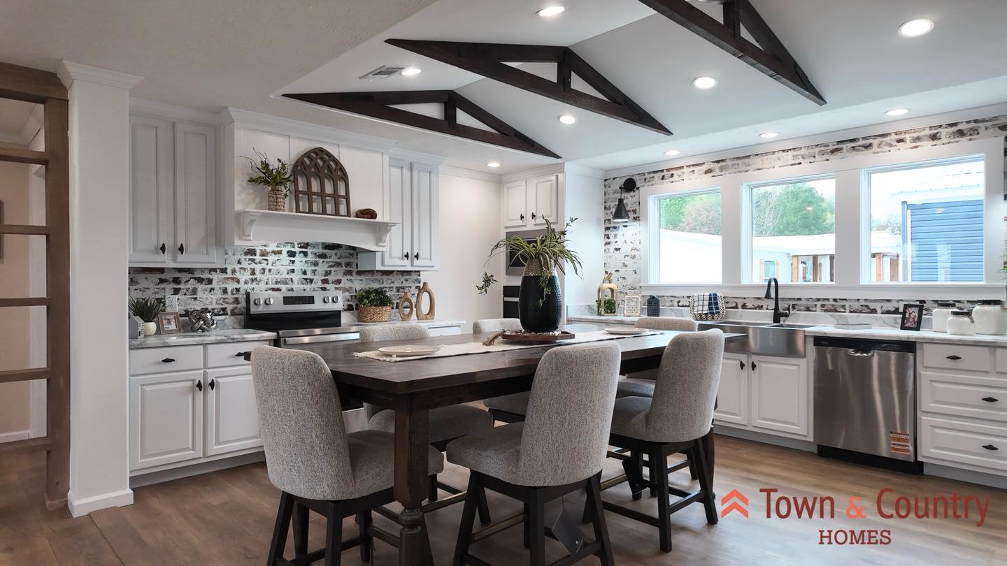 A modern kitchen with a central dining table, four upholstered chairs, and wooden beam ceiling accents. White cabinets, stainless steel appliances, and large windows create a bright, inviting atmosphere.