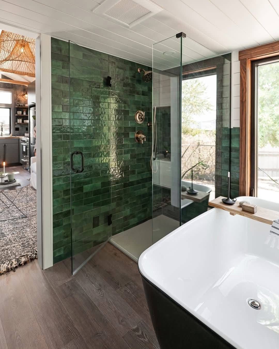 Modern bathroom with sleek glass shower, featuring deep green tiles and gold fixtures. A freestanding bathtub and large window add a bright, airy touch.