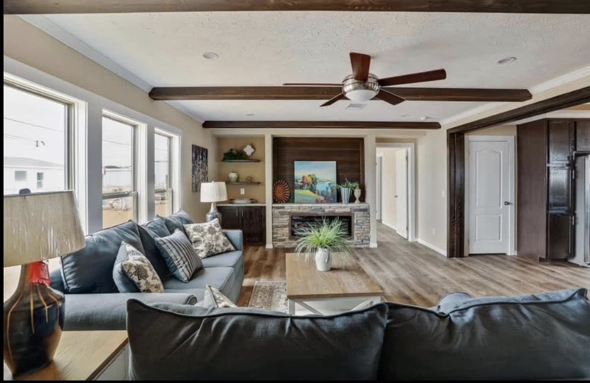 Modern living room with wooden floors and ceiling beams, a ceiling fan, large windows, gray sofas with patterned cushions, and a fireplace with artwork. Cozy and inviting atmosphere.