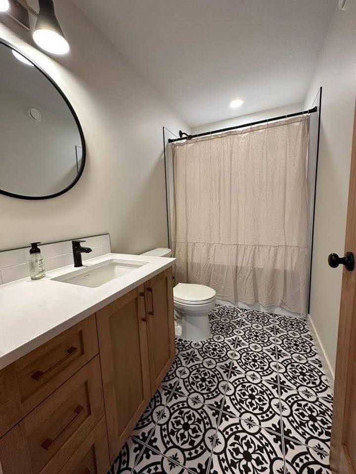 Modern bathroom with geometric black and white floor tiles, a wooden vanity with a white countertop, and an oval mirror. A shower curtain hangs in the background.