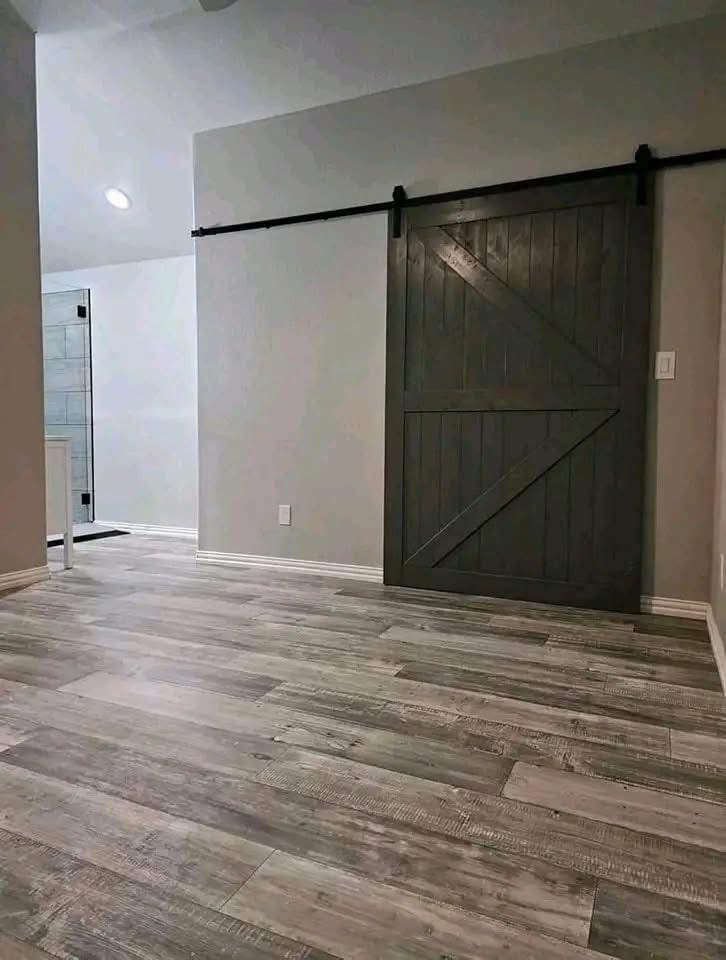 A modern room features light wood flooring and a dark wooden barn door on a track against a grey wall. The space appears clean and minimalist.