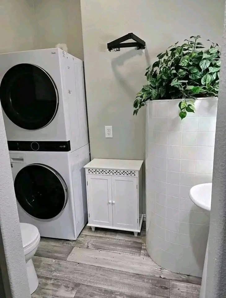 Laundry and bathroom hybrid space with stacked washer and dryer, white cabinet, curved tiled divider with lush greenery, and toilet on wooden floor.