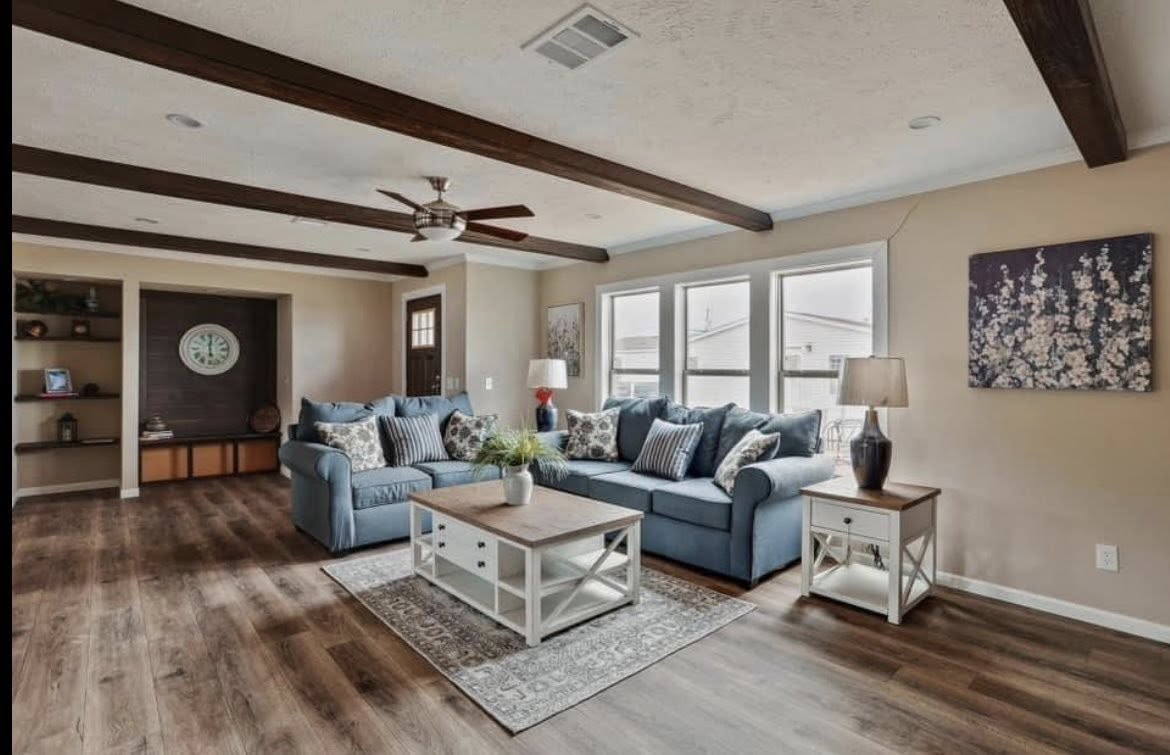 Spacious living room with blue sofas, wooden beams, and large windows. A cozy ambiance with decorative pillows, a rug, and a ceiling fan.