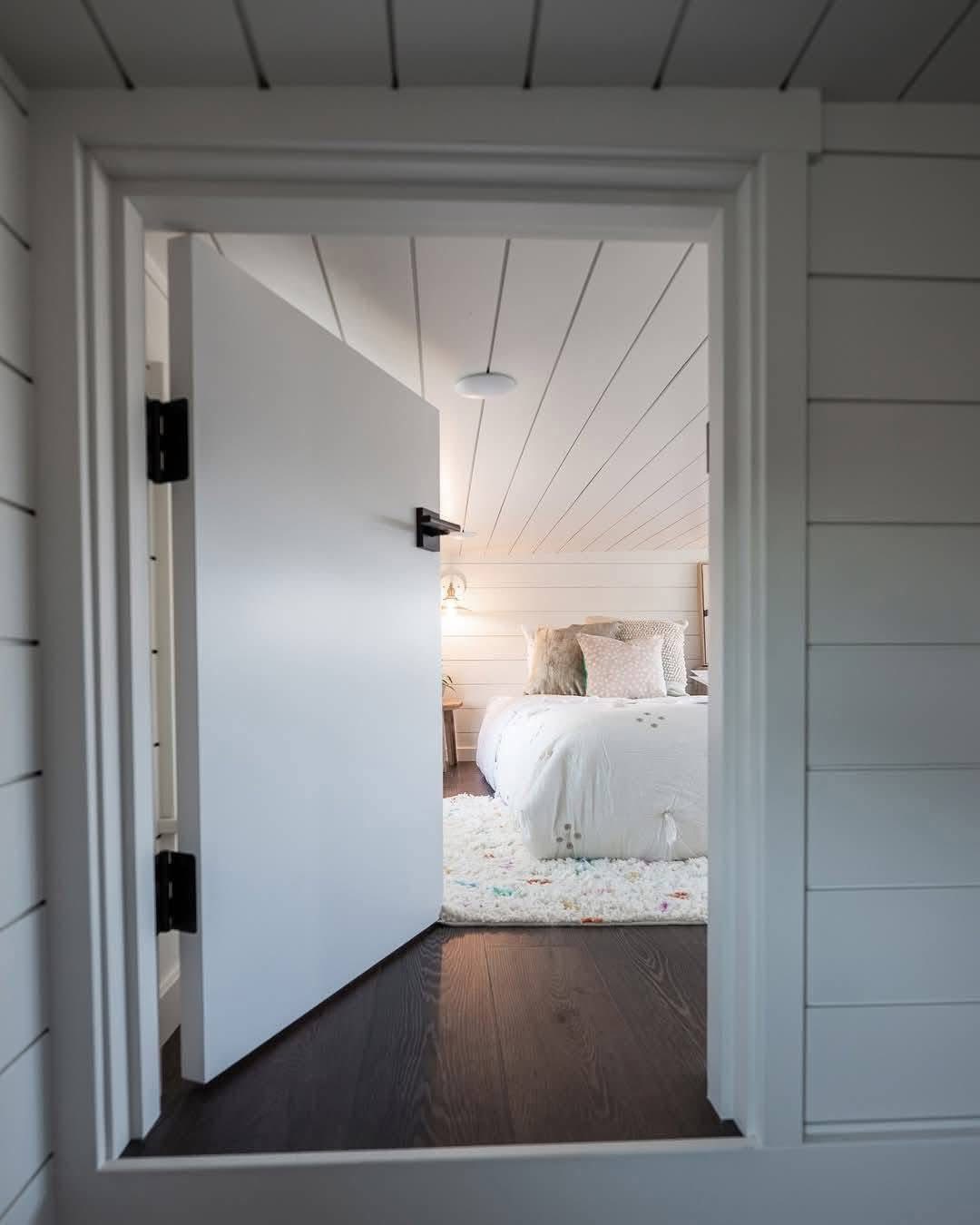 View through a partially open white door into a cozy bedroom with white shiplap walls, soft bedding, a plush rug, and a warm, inviting atmosphere.