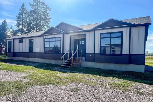 A modern modular home with large windows and a gabled roof sits on a grassy area under a clear blue sky. Stairs lead to a central entrance.