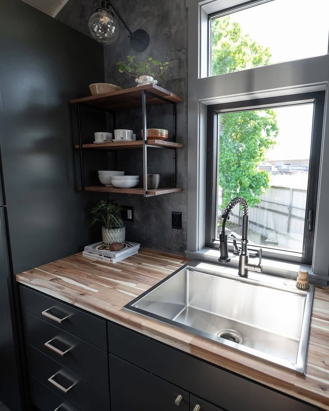 A modern kitchen corner with gray cabinets and a wooden countertop. Open shelves hold dishes beside a stainless steel sink. A window shows lush greenery.