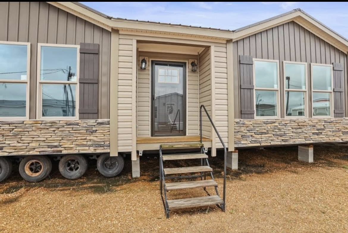 A beige modular home with stone accents and six windows. It sits on wheels and has a glass door with steps leading up, set against a clear sky.