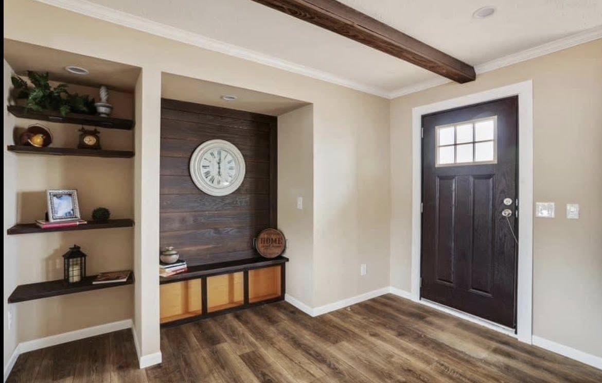 Cozy entryway with wood accents. A dark wooden door is on the right. Shelves with decor and a clock adorn the left wall, creating a warm and inviting tone.