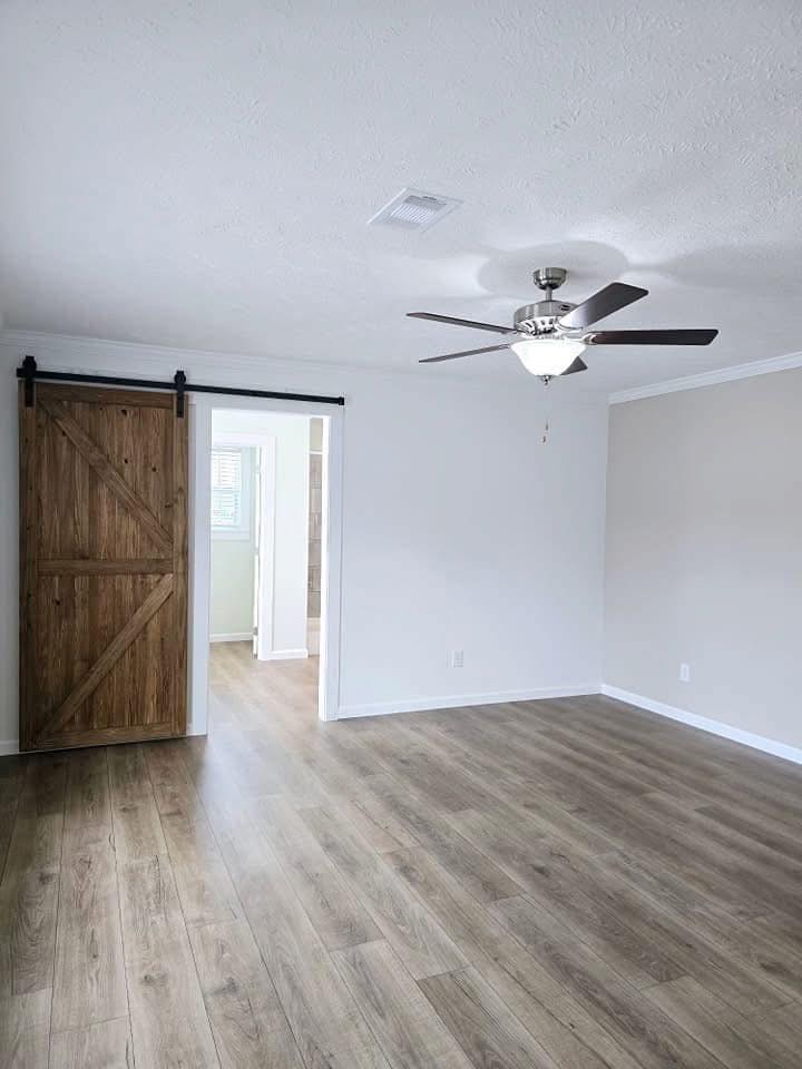 Spacious room with light wooden flooring, a ceiling fan, and a sliding barn door leading to a bright adjoining room. Neutral walls create a serene tone.