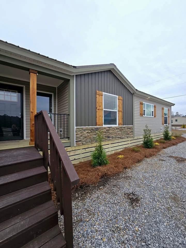 Single-story, modern house with gray siding and stone accents. Wooden shutters frame large windows. A brown staircase leads to a porch. Sparse landscaping includes small shrubs.