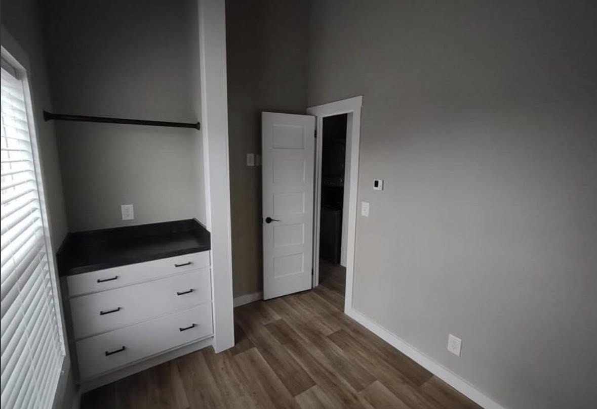 A small room with light gray walls, wood flooring, and a white door slightly ajar. On the left, there's a built-in cabinet with three drawers and a black countertop beneath an open closet bar. A window with blinds is partially visible. The room feels minimal and clean.