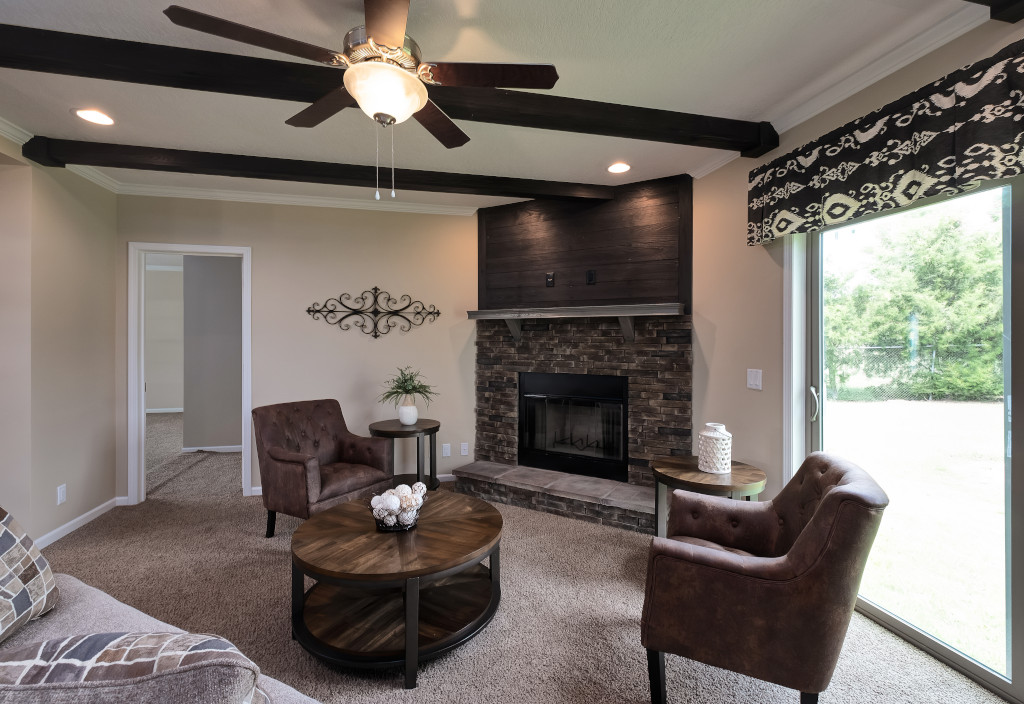 Cozy living room with a fireplace, two brown armchairs, and a round wooden coffee table. Ceiling fan and beams add warmth. Large window with patterned valance.