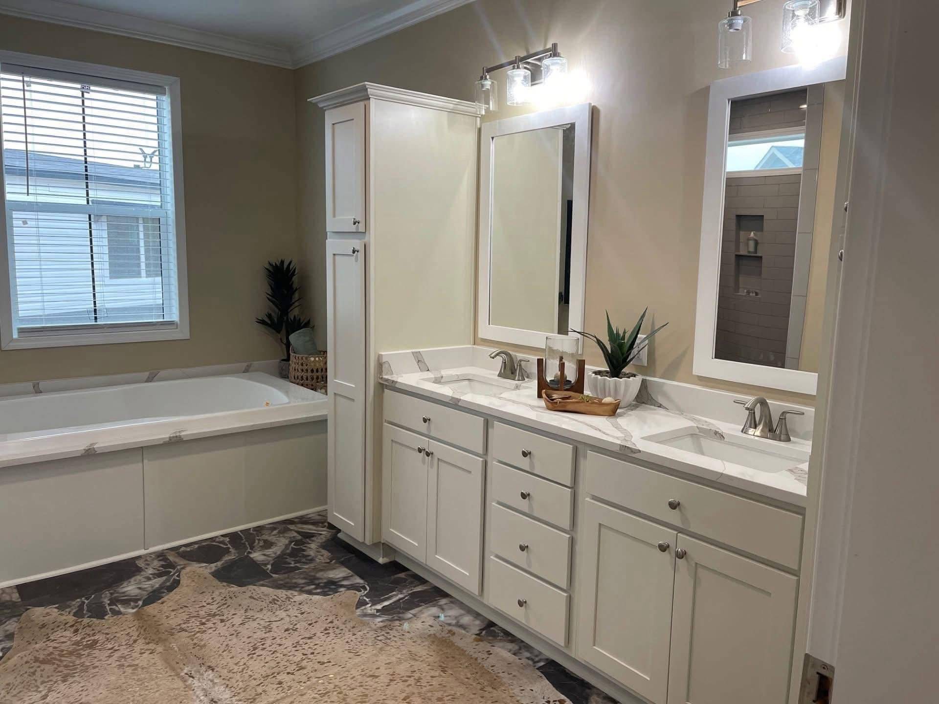 Modern bathroom with cream walls and a dark marble floor. Features a dual-sink vanity with mirrors and lights, a large tub by a window, and plants for decor.