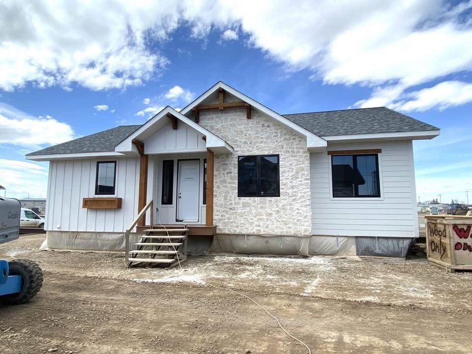 Small modern house under construction with a white stone facade and a gabled roof. A wooden staircase leads to the front door. Bright blue sky above.