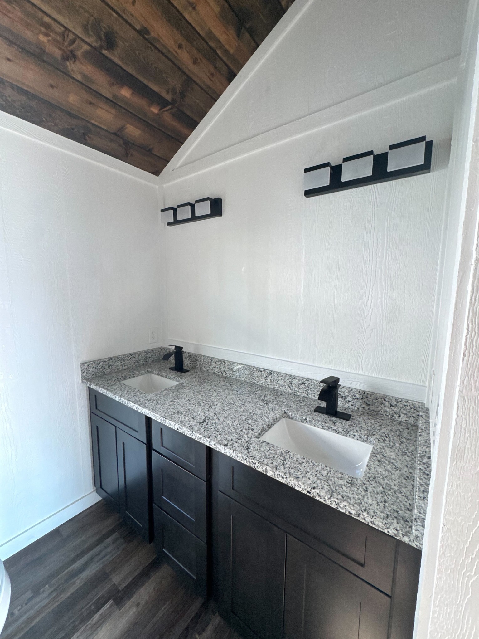 Elegant bathroom with a vaulted wood ceiling, dual granite sinks, and dark wood cabinets. Modern black fixtures and bright, clean ambiance.