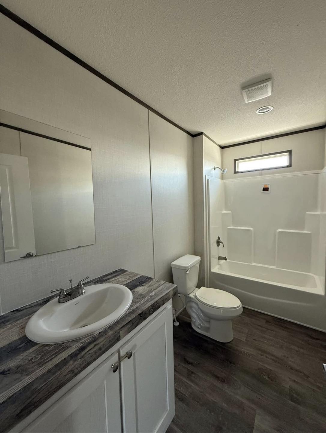 Modern bathroom with a white vanity and sink, wood-patterned countertop, wall mirror, toilet, and shower-tub combo under a small window, evoking a clean, minimalistic feel.