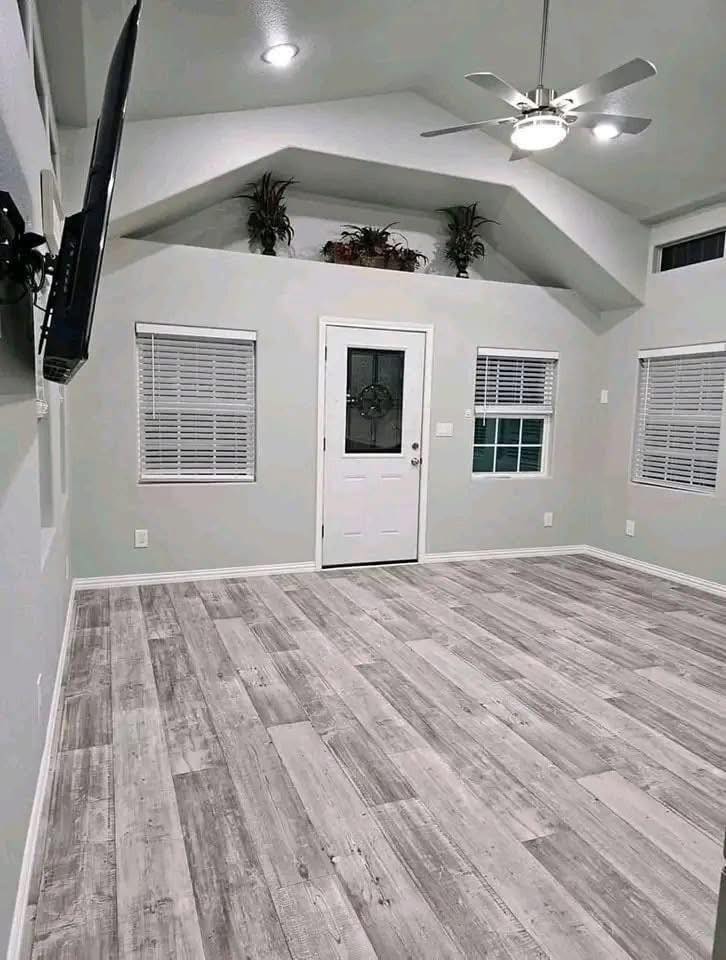 Modern room with light gray walls and wood floor. Features a white door, two windows, ceiling fan, wall-mounted TV, and plants on a high shelf.