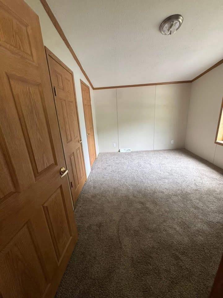 Empty room with beige carpet and cream walls. Two wooden doors are on the left, a small window on the right, and a ceiling light fixture. Simple and clean.