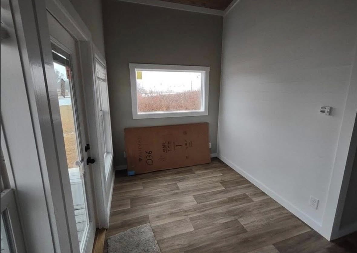 Small, unfurnished room with light wood flooring, white walls, and a large window revealing a cloudy, overcast sky. A cardboard box leans against the wall, suggesting recent move-in or renovation.