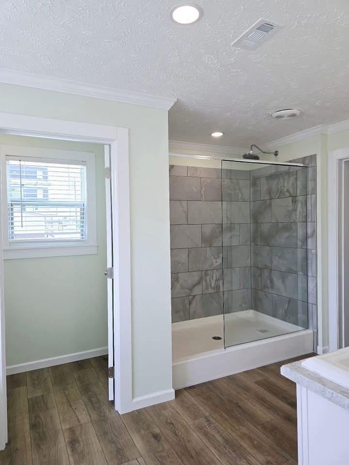 Bathroom with a walk-in shower enclosed by glass, featuring large gray tiles. Light green walls, wooden floor, and a window with blinds create a serene atmosphere.