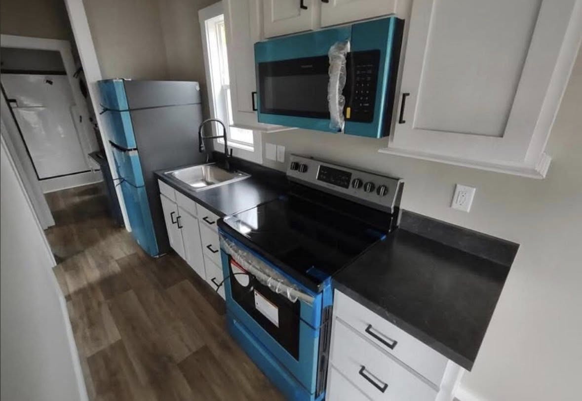 Modern kitchen with blue-accented fridge, stove, and microwave. Black countertops, white cabinets, and wood flooring create a sleek, minimalist look.