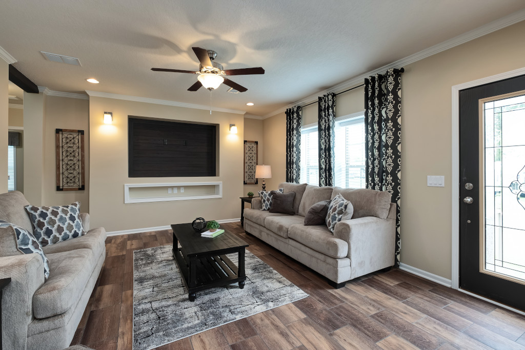 A modern living room with beige walls, hardwood floors, a ceiling fan, and two beige sofas with patterned cushions. Large window with black-and-white curtains on the right.