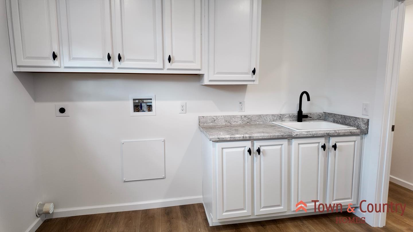 A laundry room with white cabinets, a gray countertop, and a black faucet over the sink. The wood floor adds warmth to the clean, bright space.