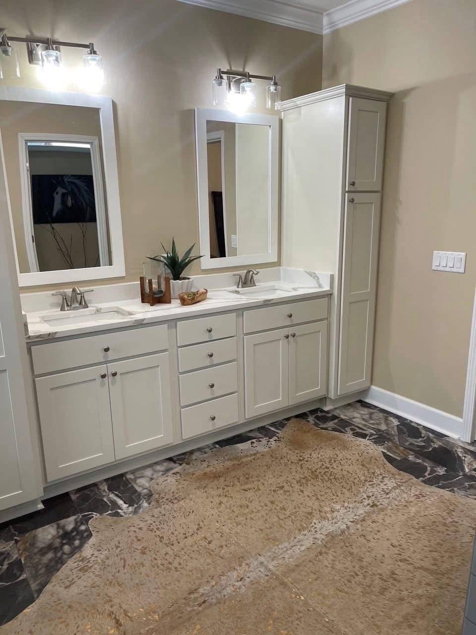 Elegant bathroom with a double sink vanity and marble countertops. Two illuminated mirrors are above each sink. A cowhide rug on marble flooring adds warmth. Neutral tones create a calming ambiance.
