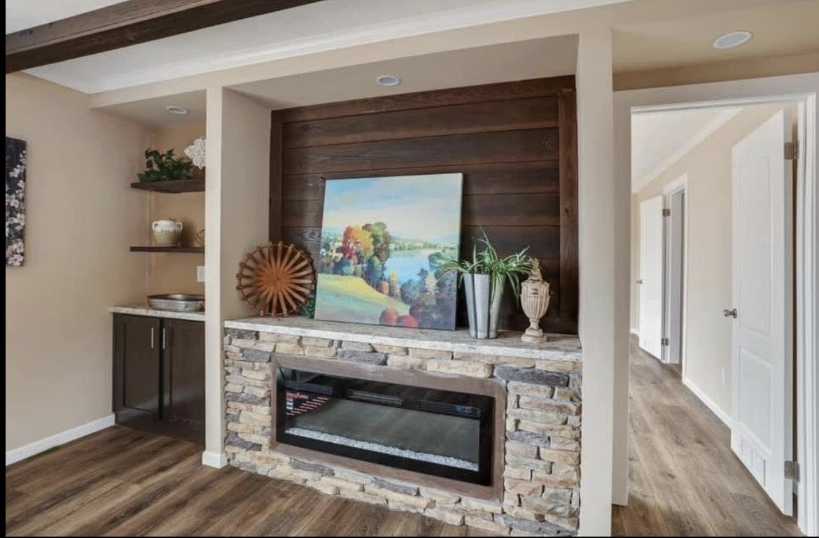 Cozy living room with a stone-framed electric fireplace beneath a vibrant landscape painting. Adjacent shelves hold decor items, creating a warm, inviting atmosphere.