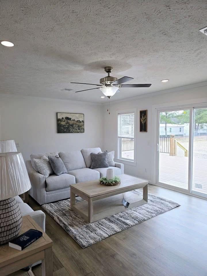 A bright living room with a light gray sofa and cushions, a wooden coffee table on a striped rug, and a ceiling fan. Large windows overlook a deck.