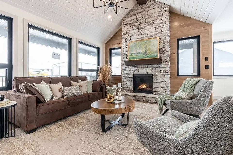 Cozy living room with a brown sofa, gray armchairs, and a wooden coffee table. A stone fireplace with a landscape painting and large windows enhance the airy, warm ambiance.