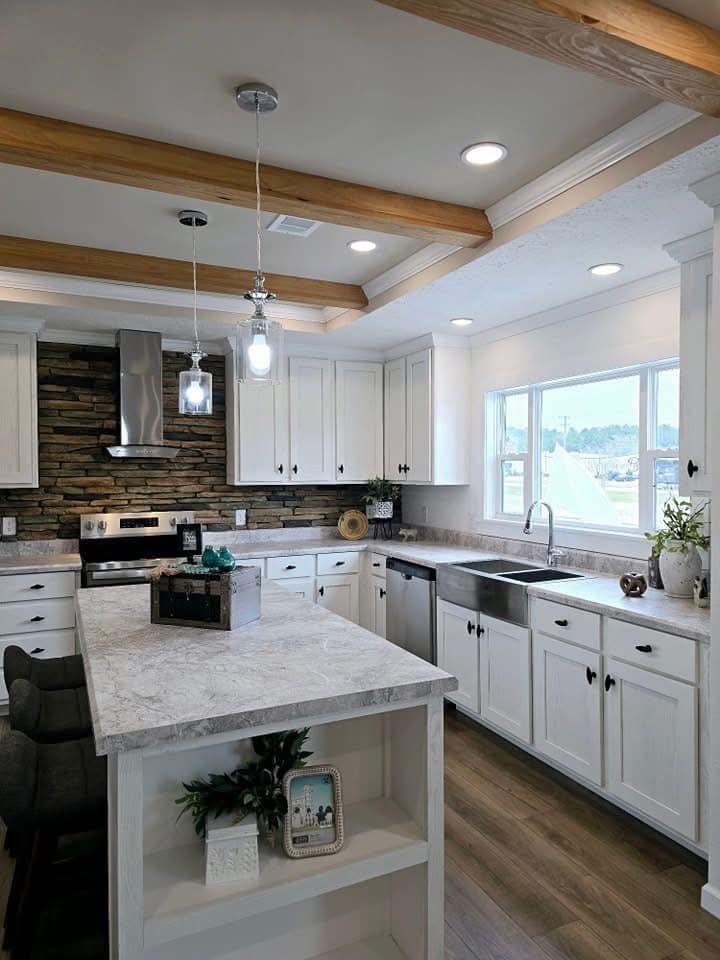 Modern kitchen with white cabinetry and a stone backsplash. Features a marble island, stainless steel appliances, and wooden ceiling beams. Bright natural light from a large window adds warmth.