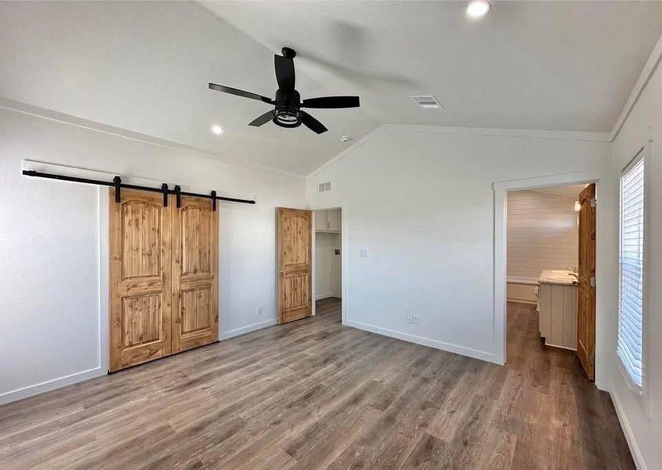 Spacious room with wooden sliding barn doors, light wood flooring, ceiling fan, and white walls. Bright natural light from large windows creates an airy feel.