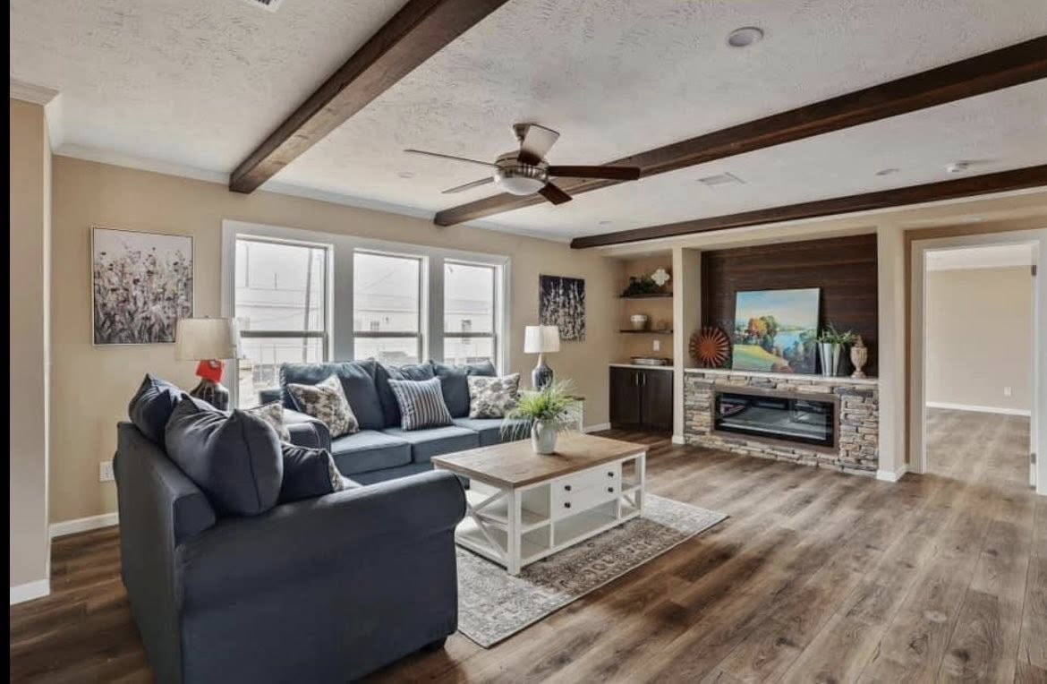 A cozy living room with a navy blue sectional sofa, wooden coffee table, and a stone fireplace. Large windows, ceiling beams, and neutral tones create warmth.